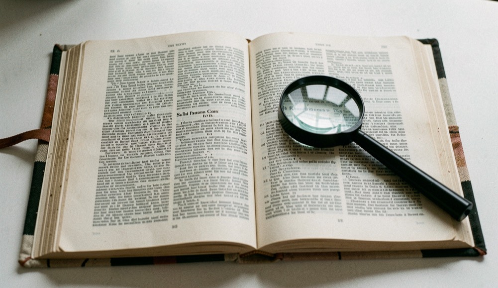 A cloth bound dictionary open on a dense double page, a black handled magnifier lying across the right hand column