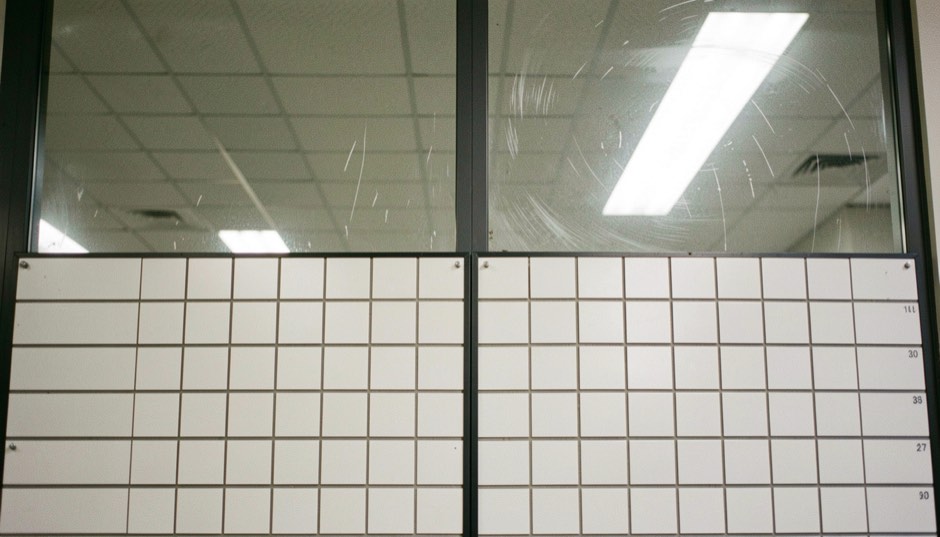 A split flap departure board of blank white tiles behind scratched glass, reflecting a pale ceiling of strip lights